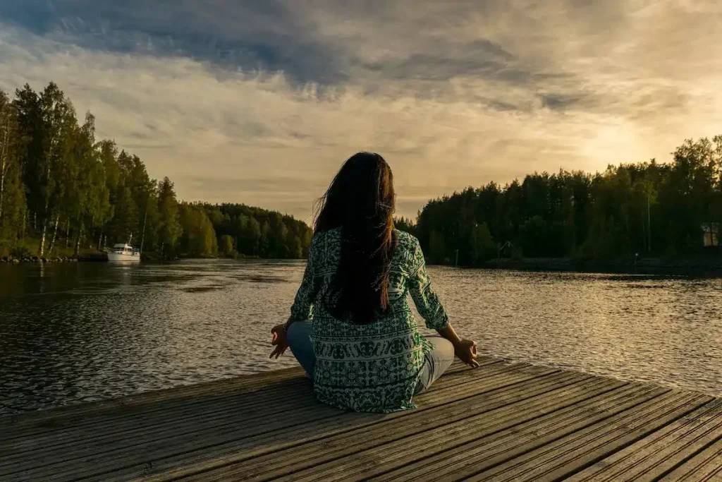 Person meditating on a wooden dock by a calm lake surrounded by forest at sunset