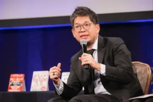 Steve Chen speaking on stage at a conference while holding a microphone and wearing a black suit.