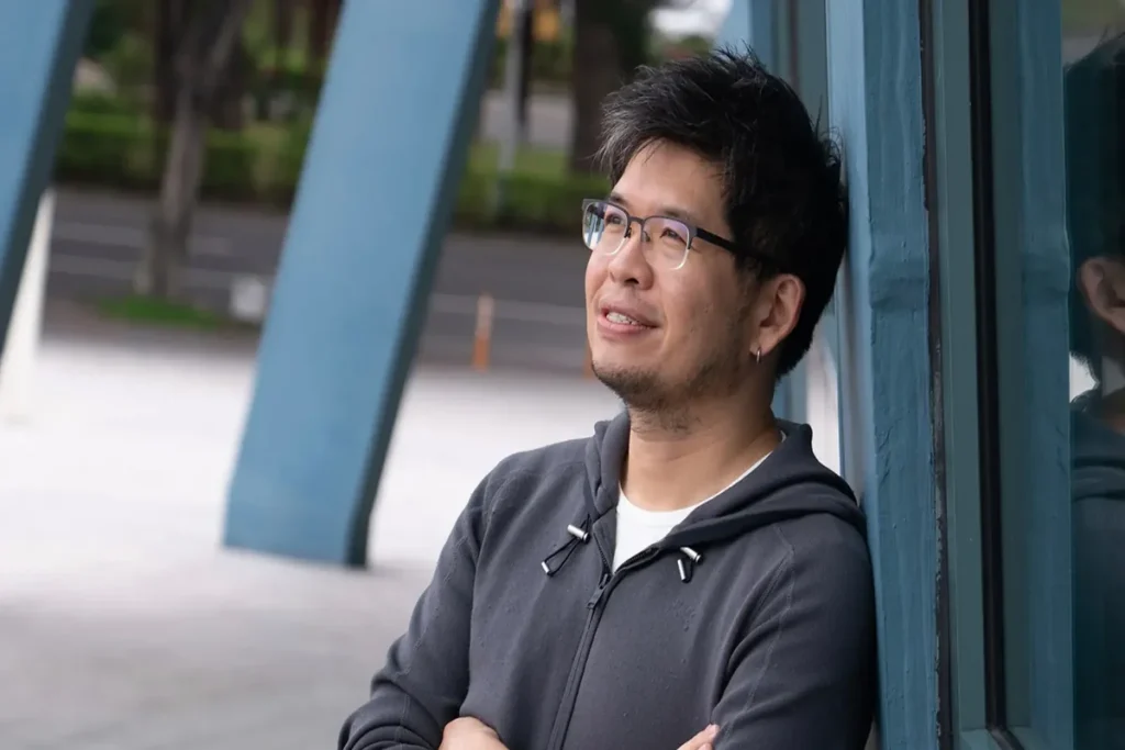 Steve Chen leaning against a blue wall outdoors, wearing glasses and a dark hoodie while looking thoughtfully into the distance.