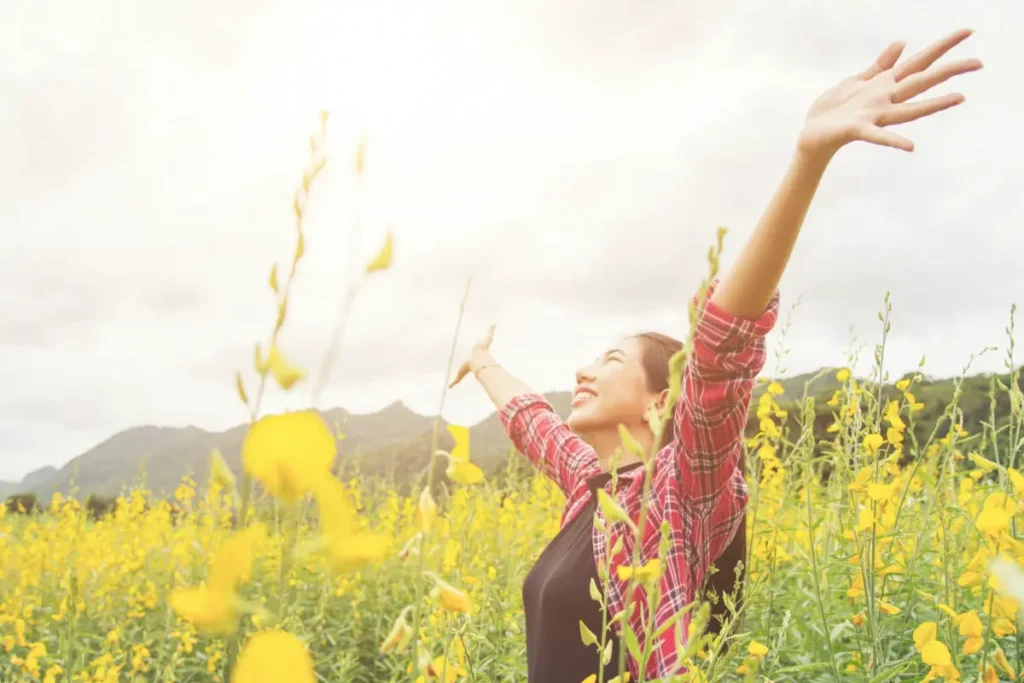 Woman with arms raised in a sunlit field of yellow flowers expressing joy and well-being - happiness and high performance keynote | Aurum Speakers Bureau